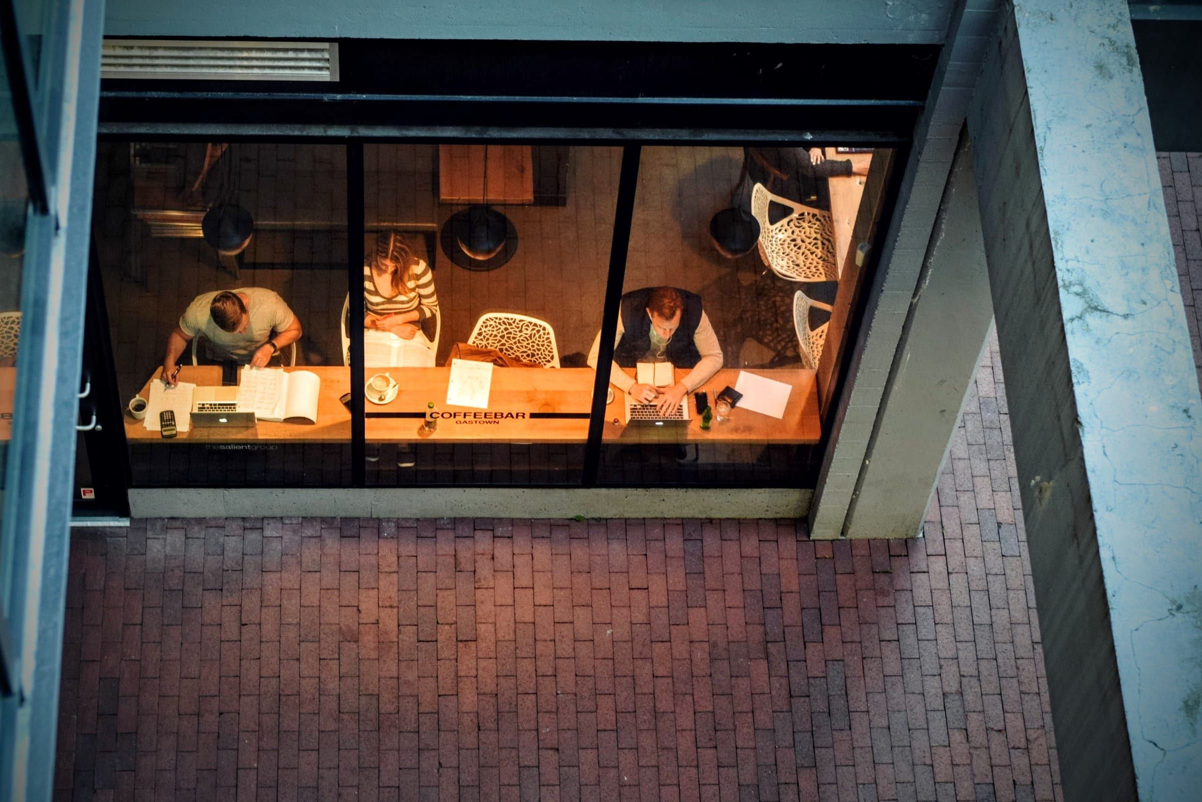Seen from above at dusk: three people working with laptops and notebooks at a coffee bar window.