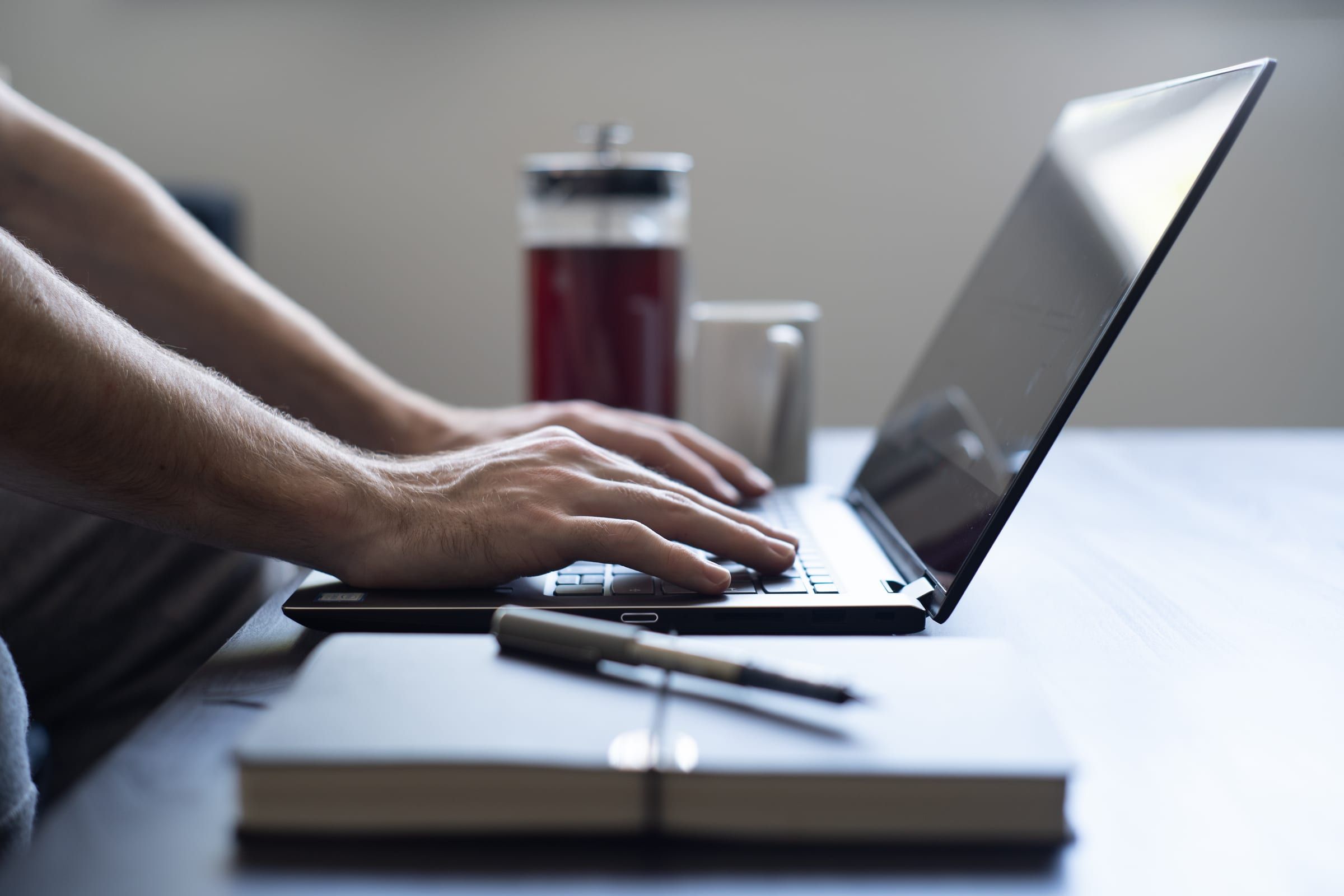 Hands typing on a laptop at a bright table, a notepad with a pen and a glass bottle of drink beside it.